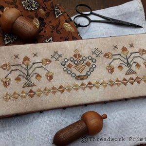 May include: A rectangular, beige cross-stitch sampler with acorn and floral patterns in shades of brown and gold. Two wooden acorn ornaments and a pair of black scissors are also visible. The sampler is on a white and brown patterned fabric.