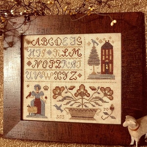 May include: A framed cross-stitch sampler with a brown wooden frame. The sampler features a brown and white alphabet, a small house with a tree, a potted plant with flowers, and a woman with a lamb. The sampler is on a brown and gold patterned surface.