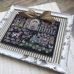 May include: A framed cross-stitch design featuring a farmhouse scene with a red barn, a blue truck, cows, chickens, and beehives. The design is stitched on black fabric with colorful thread.