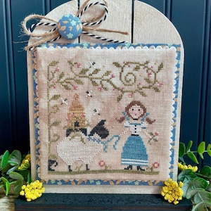 May include: A framed cross-stitch featuring a sheep and a girl in a blue dress. The cross-stitch is in a wooden frame with a blue and white patterned bow at the top. The cross-stitch has a beige background with green and pink accents.