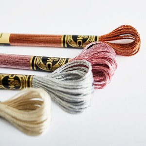 May include: Four spools of DMC embroidery floss in shades of copper, rose gold, silver, and cream. The spools are labeled with the DMC brand name and the color number.