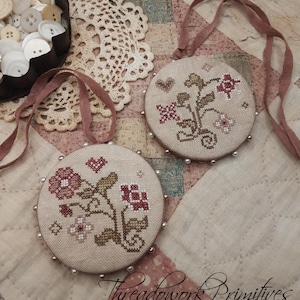 May include: Two round, beige, embroidered ornaments with a floral design in shades of red, pink, and green. Each ornament has a brown ribbon attached and is surrounded by small, white beads.