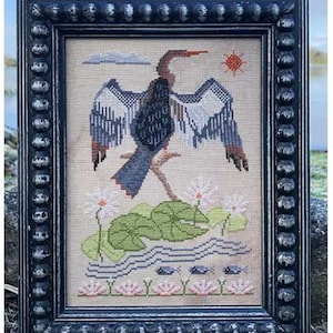 May include: A cross-stitch embroidery of a black bird with outstretched wings standing on lily pads in a pond. The bird is surrounded by water lilies and a blue sky with a white cloud. The embroidery is framed in a black frame with a decorative border.