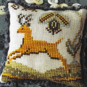May include: A beige pillow with a gray border features a cross-stitch design of a yellow deer jumping over a green bush. The deer has large antlers and a long tail. The background is a light beige with a green and brown floral pattern.