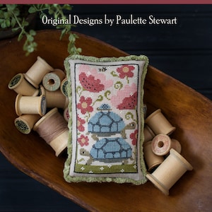 May include: A cross-stitch sampler pillow with a green border and a design of three tortoises stacked on top of each other, surrounded by pink flowers. The pillow is sitting in a wooden bowl with spools of thread. The text "Tortoise Tower" is at the bottom of the image.