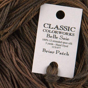 May include: Close-up of brown yarn with a white tag. The tag reads "CLASSIC COLORWORKS Belle Soie" and "Briar Patch." The yarn is 100% 12-stranded spun silk, hand-dyed, and 5 yards in length. The color is a mix of brown shades.