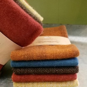 May include: A stack of colorful wool fabric squares in shades of red, orange, blue, and brown. The fabric has a textured, felted appearance. The stack is bound with a paper band.