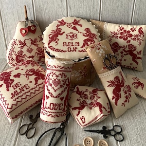May include: A collection of handmade Valentine's Day themed needlepoint pillows and a round box with a lid. The pillows feature red and white embroidery with cherubs, hearts, and the words "Be Mine", "Love", and "Rely On".