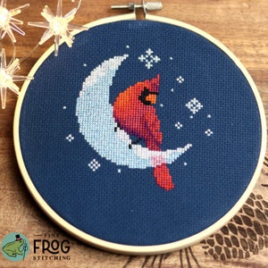 May include: Cross-stitch embroidery in a wooden hoop features a red cardinal bird perched on a crescent moon against a dark blue background. White stars are scattered around the moon. The text "Fine Frog Stitching" is at the bottom.