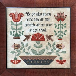 May include: Framed cross-stitch artwork featuring the text "Be ye also ready. The son of man cometh at an hour ye not think." The design includes floral and bird motifs in red, green, and brown, set against a cream background. The frame is made of dark wood.