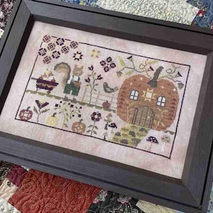May include: Framed cross-stitch art featuring a whimsical autumn scene. A squirrel and a pumpkin house are central, surrounded by flowers, a bird, and pumpkins. The artwork is framed in a dark brown wooden frame, set against a colorful quilt.