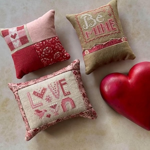 May include: A collection of decorative pillows and a heart-shaped object. The pillows feature cross-stitch designs with the words "Be Mine" and "LOVE", along with heart and floral patterns. The heart is a solid, glossy red.
