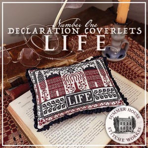 May include: A decorative pillow with a cross-stitch design featuring two red houses, a weeping willow, and the word "LIFE." The pillow has a black ruffled edge and is set on an open book. The text "Number One DECLARATION COVERLETS LIFE" is also visible.