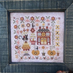 Peut inclure: Une broderie au point de croix encadrée dans un cadre en bois vert foncé. Le motif représente une maison rouge avec une porte et des fenêtres jaunes, deux chats noirs, des citrouilles et le texte "Happy Autumn".