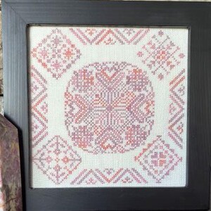 May include: Square framed cross-stitch art with a geometric pattern in shades of pink, peach, and lavender. The central design is a large, detailed circular motif, surrounded by smaller square and triangular patterns. The artwork is set against a white background within a black frame.