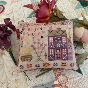 May include: A cross-stitch sampler with a pink and purple house and the text "sampler house" in a cream colored frame. The sampler is on a white and pink quilt with a pair of rose gold scissors.