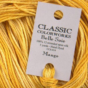 May include: Close-up of a skein of yellow Classic Colorworks Belle Soie thread. The label reads "Mango" and details the thread as 100% 12-stranded spun silk, 5 yards, hand dyed. The thread is a vibrant yellow with lighter yellow highlights.