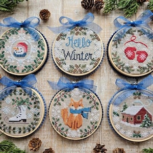 May include: Six embroidered winter-themed ornaments. Each round design features a different image: a snowman, the text "Hello Winter", red mittens, an ice skate, a fox, and a snow-covered house. Each ornament has a blue ribbon.