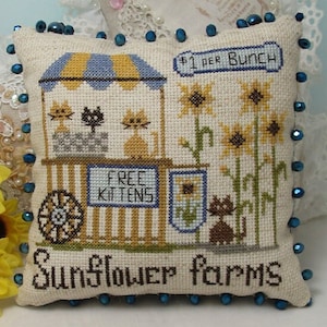May include: A decorative pillow with a cross-stitch design. The design features a sunflower stand with kittens, sunflowers, and the text "FREE KITTENS" and "Sunflower Farms". The pillow is trimmed with blue beads.