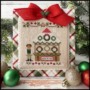 May include: Cross-stitch artwork featuring a Christmas market scene with wreaths, a red roof, and a figure. The artwork is framed with a red bow and plaid fabric. Green and gold ornaments are in the foreground.
