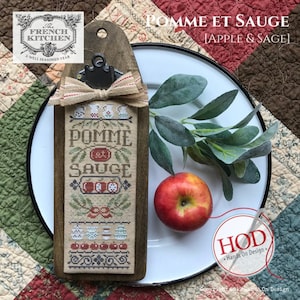 May include: A cross-stitch pattern on a wooden plaque with a brown background. The pattern features a green, red, and white design with the text "Pomme et Sauge" and "Apple & Sage".