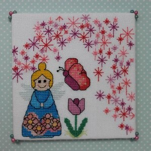 May include: Cross-stitch embroidery of an angel with pink flowers, a butterfly, and a pink tulip on a white background. The angel is wearing a blue dress with pink flowers and has blonde hair.