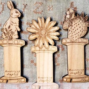 May include: Three wooden spool-shaped ornaments with different designs. The first ornament features a rabbit with flowers, the second a daisy, and the third a strawberry. Each ornament has the text "Heartstring Simply" on the bottom.