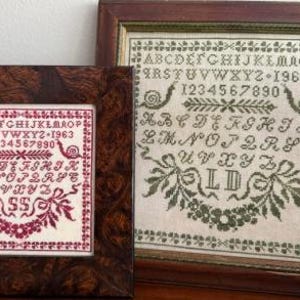 May include: Two framed cross-stitch samplers. One is framed in a dark wood with a reddish-pink design, including the alphabet, numbers, and the year 1963. The other is framed in a brown wood with a green design, including the alphabet, numbers, and the year 1960.