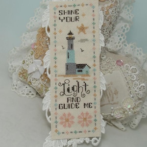 May include: A cross-stitched bookmark featuring a lighthouse design with the text "SHINE YOUR LIGHT AND GUIDE ME." The lighthouse is blue and gray, with a star and two birds. The bookmark has a white lace border and floral accents.