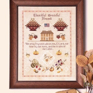 May include: A framed cross-stitch sampler with a beige background and brown frame. The sampler features a house, a basket of fruit, a pumpkin, and the text "Thankful Grateful Blessed. We pray Thy Love Willbless, O Lord, our hearts, our home, and the Fruits of our Labor."
