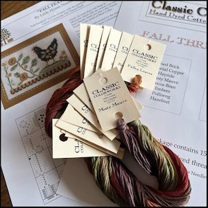 May include: A collection of hand-dyed cotton embroidery floss in shades of brown, green, and purple, with tags from Classic Colorworks. The image includes a cross-stitch pattern with a chicken design and the text "Fall Feathers."