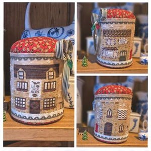 May include: A collection of three decorative pincushions with a Christmas theme. Each pincushion is cylindrical, featuring cross-stitch designs of buildings with windows and doors. The tops are red with a leaf pattern, and they are adorned with striped ribbons.