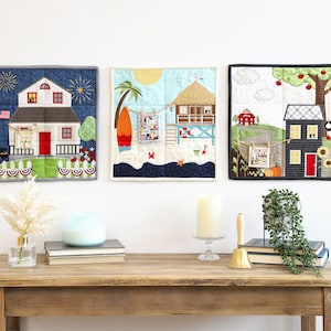 May include: Three framed fabric art pieces depicting houses and beach scenes. The first shows a house with fireworks, the second a beach scene with a surfboard, and the third a house with a tree and pumpkins.