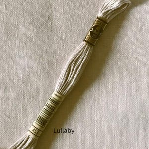 May include: A skein of DMC 25 embroidery floss in a light beige color, labeled "Lullaby" and marked with the number 453. The floss is tightly wound and wrapped with a paper band featuring the DMC logo and product information. The background is a textured, off-white fabric.
