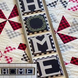 May include: A framed wooden sign with the word "HOME" spelled out in black and white letters. Each letter is on a separate square canvas with a black and white checkered border. The sign is hanging on a white and black checkered quilt.