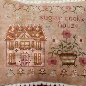 May include: A cross-stitch pillow featuring a gingerbread house with a brown roof and white trim. The house is surrounded by flowers and trees. The text "sugar cookie house" is stitched above the house.