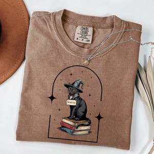 May include: A brown crewneck t-shirt featuring a graphic of a black cat wearing a witch hat, sitting on a stack of books with a sign that reads "Read me". The design is framed by a black arch and stars, with a silver necklace resting on the collar.