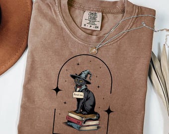 Könnte beinhalten: Ein braunes T-Shirt mit Rundhalsausschnitt und einem Aufdruck einer schwarzen Katze mit Hexenhut, die auf einem Stapel Bücher sitzt und ein Schild mit der Aufschrift "Read me" hält. Das Motiv ist von einem schwarzen Bogen und Sternen umrahmt.