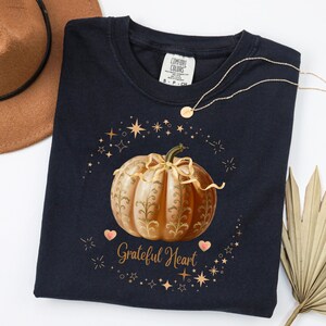 May include: A navy blue t-shirt featuring a detailed illustration of a pumpkin with gold accents and the words "Grateful Heart". The design is surrounded by stars and hearts. A gold necklace is draped across the shirt.