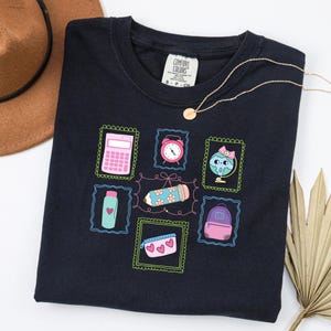May include: A navy blue t-shirt with a school-themed design. The illustrations include a pink calculator, a clock, a globe with a bow, a pencil, a water bottle, a pencil case with hearts, and a backpack. A gold necklace is draped across the shirt.