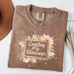 May include: A brown Comfort Colors t-shirt with the text "Counting My Blessings" in a gold frame, surrounded by floral graphics. A silver necklace with a pendant is draped across the shirt. The shirt is size S-P-CH.