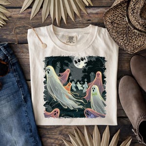May include: Cream-colored t-shirt featuring a Halloween-themed graphic. The design depicts a graveyard scene with cartoon ghosts, a full moon, and the word "BOO!" in a circular text. The ghosts are in various colors, including white, orange, and pink.