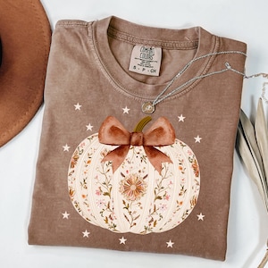 May include: A brown Comfort Colors t-shirt with a white pumpkin design, brown bow, and floral details. The shirt has a crew neck and short sleeves. A silver necklace with a pendant is draped across the shirt. Small white stars are scattered around the pumpkin.