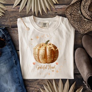 May include: Cream-colored t-shirt featuring a detailed illustration of a pumpkin with gold accents and the words "Grateful Heart." The design includes hearts and stars. The shirt is displayed with denim jeans, a necklace, and boots.