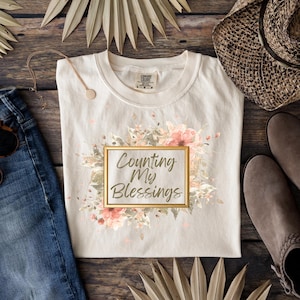 May include: Cream-colored t-shirt with the text "Counting My Blessings" inside a gold frame, surrounded by a floral design in pink and peach tones. The shirt is displayed with denim jeans, a necklace, and boots.