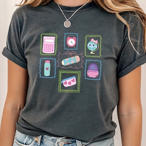 May include: Dark grey t-shirt with a school-themed design. The illustrations include a pink calculator, a pink alarm clock, a globe with a bow, a water bottle with a heart, a pencil with flowers, a pencil case with hearts, and a backpack.