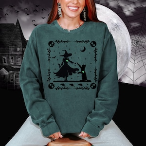 Peut inclure: Sweat-shirt vert foncé avec un motif noir d'une sorcière et d'un chat, avec une lune et des étoiles. Le motif est encadré de têtes de mort et d'éléments floraux. Vêtement confortable à manches longues.