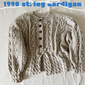 Pode incluir: Um cardigan de malha bege com um padrão de malha de cabo e um laço na cintura. O cardigan tem fecho de botões e é feito de um fio macio e quente.