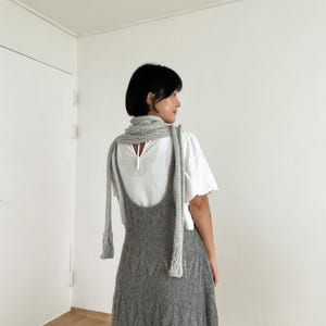 Op de afbeelding: Een grijze gebreide jurk met een lage rug en een bijpassende grijze gebreide sjaal. De jurk wordt gedragen over een witte blouse met korte mouwen en een ronde hals. De sjaal heeft een gebreid patroon met textuur en hangt langs de rug.