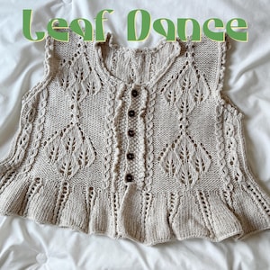 May include: A beige knitted vest with a leaf pattern and a ruffled hem. The vest has a button-up front with brown buttons and the text "Leaf Dance" in green above the vest.